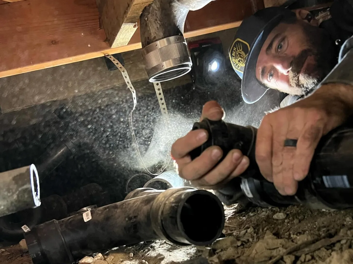 A plumber working on a residential drain services job — mid-task, hands, tools and the work itself in sharp focus, the rest of the space softly out of focus